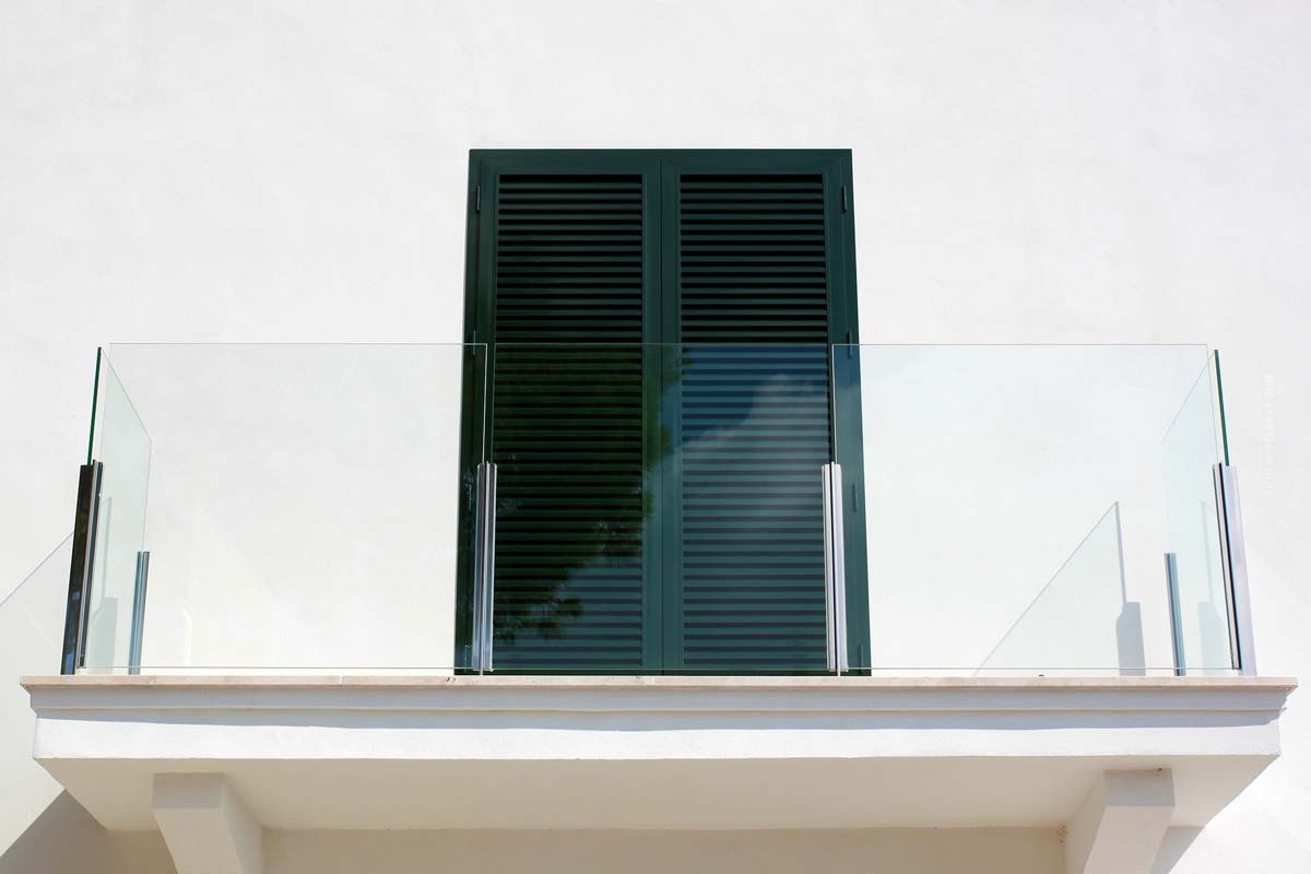 Moderner Balkon einer Neubauwohnung mit Glasgeländer und grünem Fenster an weißer Fassade